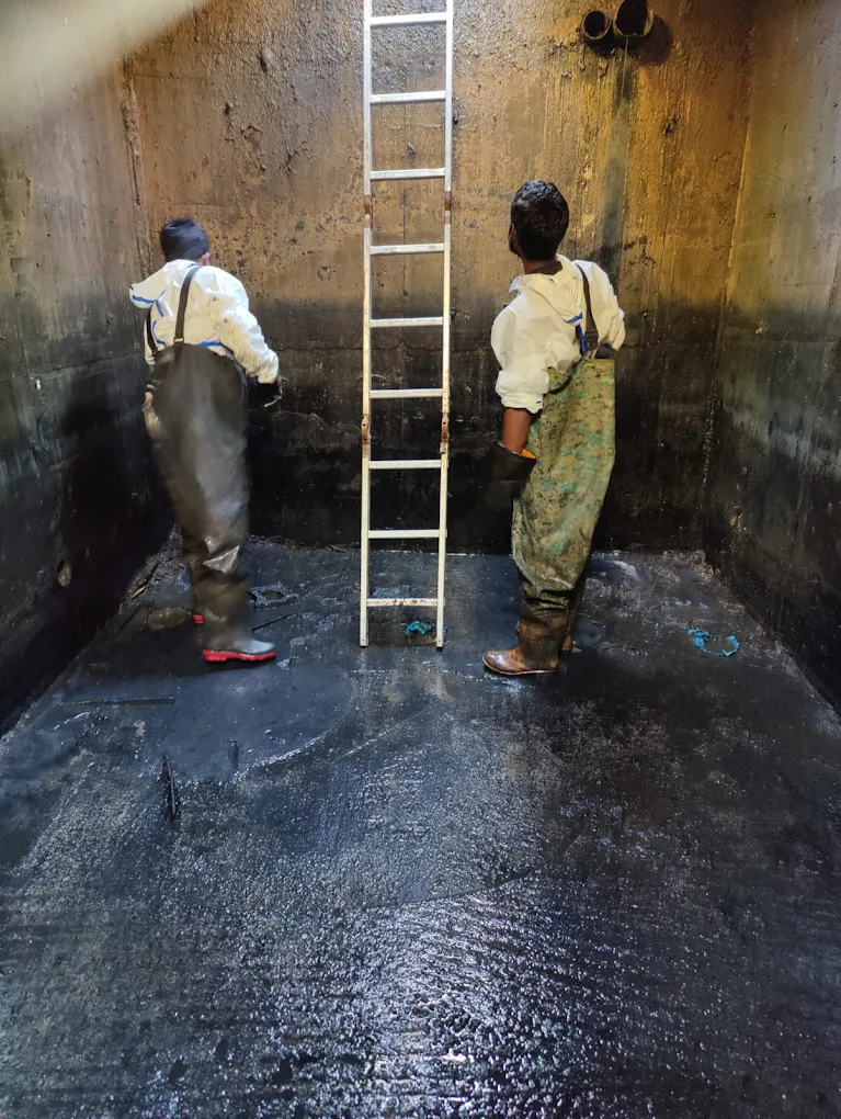 Two workers in protective waders scrubbing high walls of septic tank